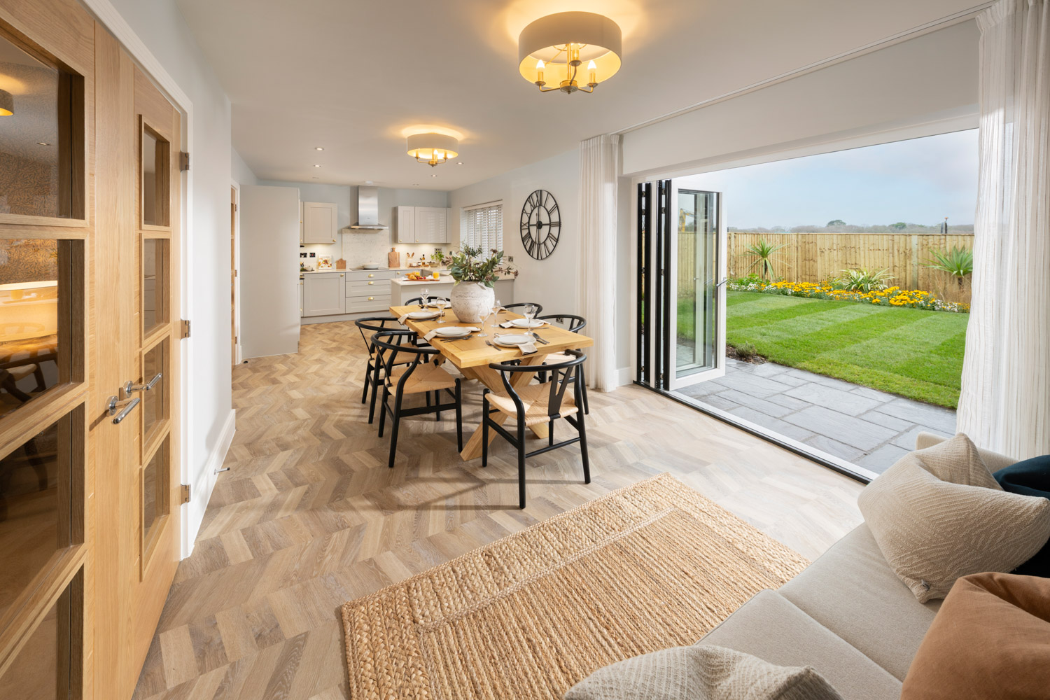 Ope plan kitchen / diner with table and chairs and wooden flooring and rug, lights on warm and homely. Bifold doors open to a green lawn with fence in background